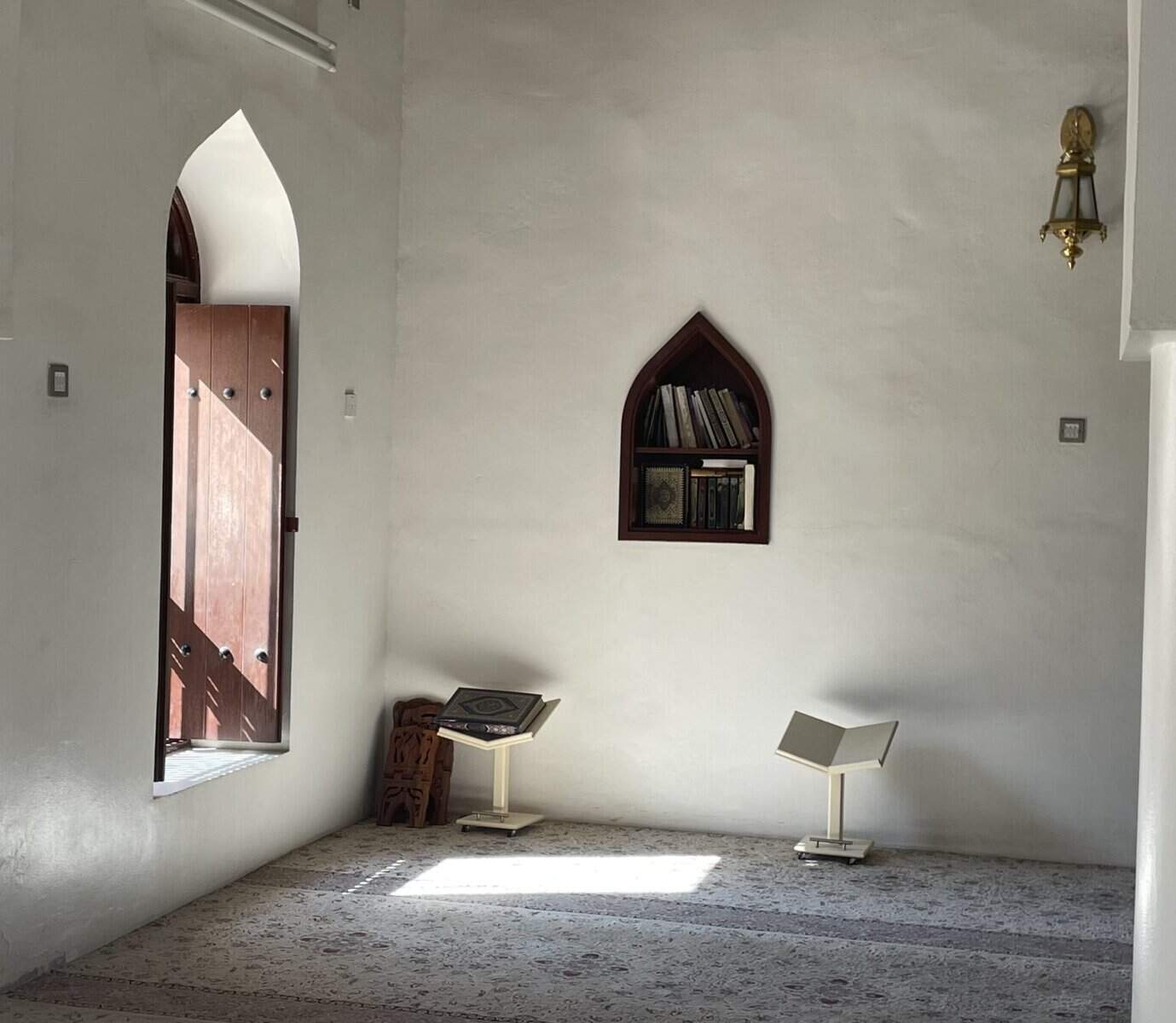 Simple interior of Al Ya’aribah Mosque with white walls, a Quran stand, and light streaming through the open wooden door.