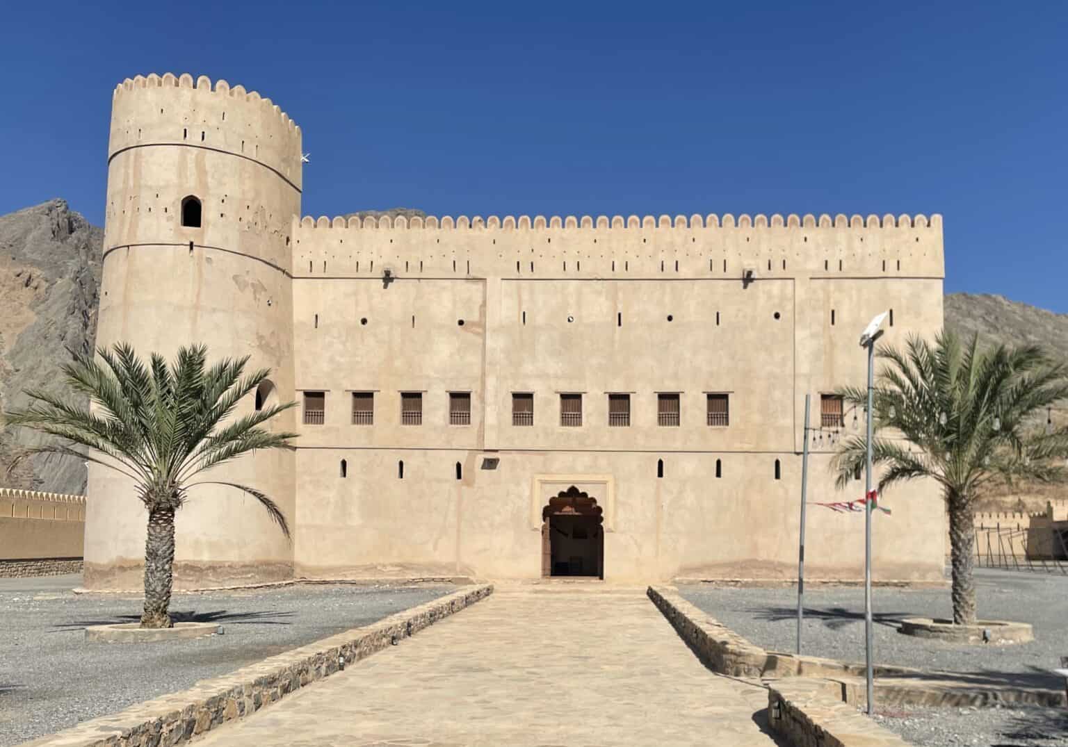 Exterior view of Bait al Ridaydah Castle in Birkat al Mouz, showing its tall round tower, fortified walls, palm trees, and the entrance pathway under a clear blue sky.