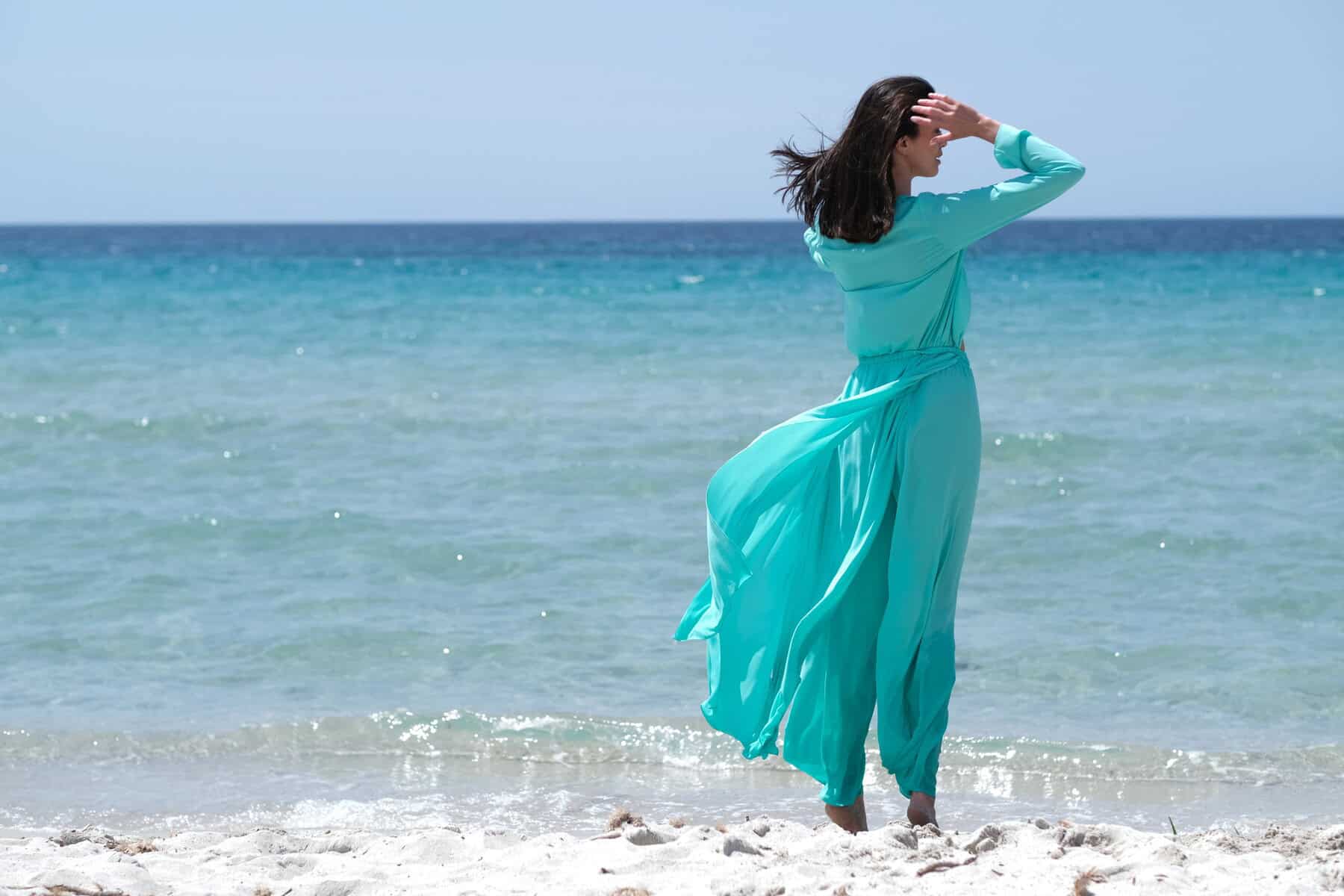 A person in a flowing turquoise dress stands on a sandy beach, facing the ocean. They hold their hair back as the sea breeze blows. Waves gently lap at the shore under a clear blue sky.