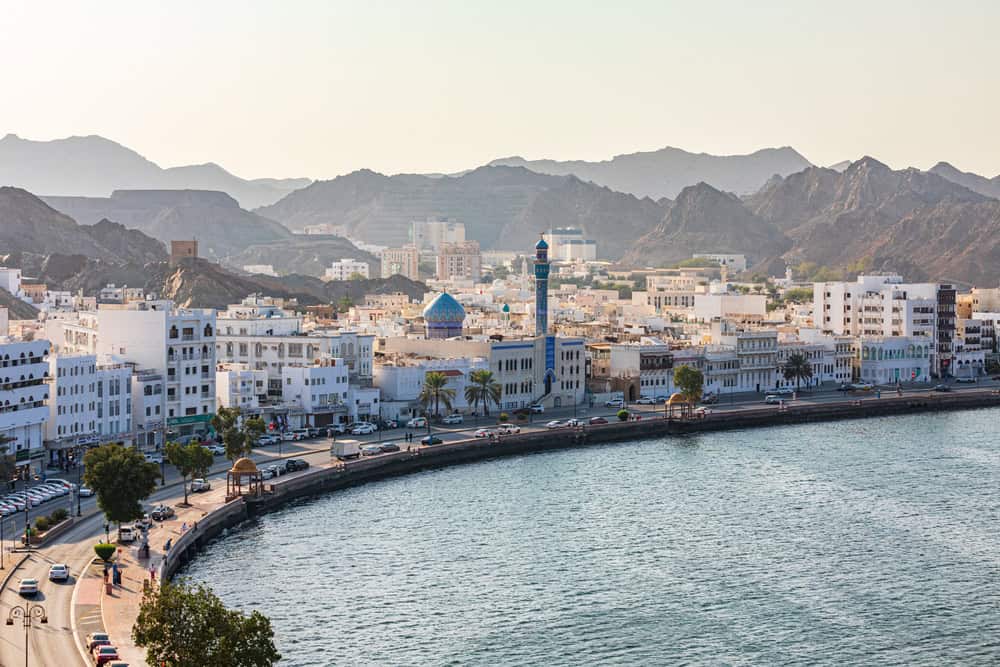 Coastal cityscape with white buildings along a curved waterfront road, bordered by a calm sea. A mosque with a blue dome and minaret stands out. Rugged mountains rise in the background under a clear sky.