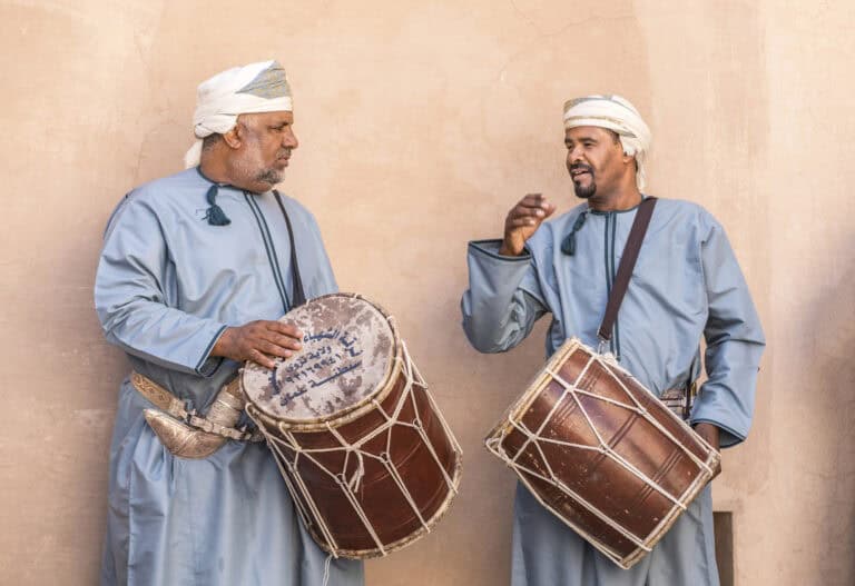 Two men in traditional attire play drums while standing against a beige wall. They wear light blue robes and white head coverings. One man gestures with his hand, as if engaged in conversation.