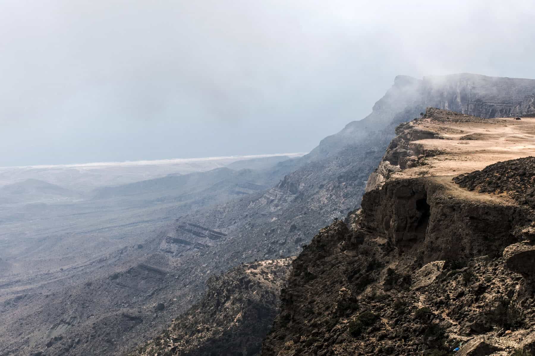A rugged, rocky cliff on the right extends into the distance under a misty sky. The landscape below features a vast, dry expanse with muted colors, fading into a distant horizon.
