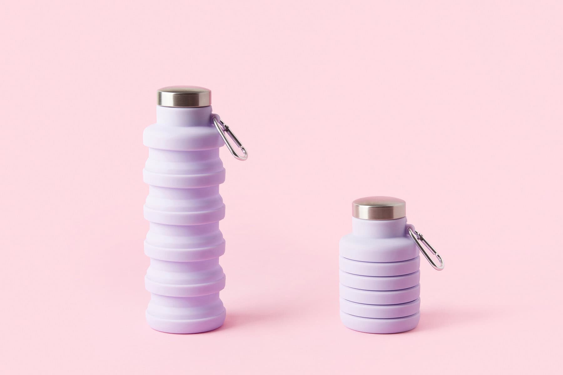 Two collapsible lavender-colored silicone water bottles with stainless steel caps and carabiners. The taller bottle is fully extended, while the shorter one is compacted. They are placed against a pastel pink background.