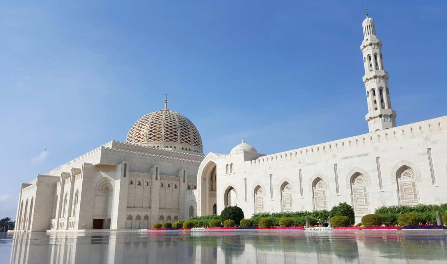 A large, white mosque with intricate carvings and a grand dome on a clear day. The structure features tall minarets and arched doorways, surrounded by manicured greenery and reflecting on a polished surface under a blue sky.