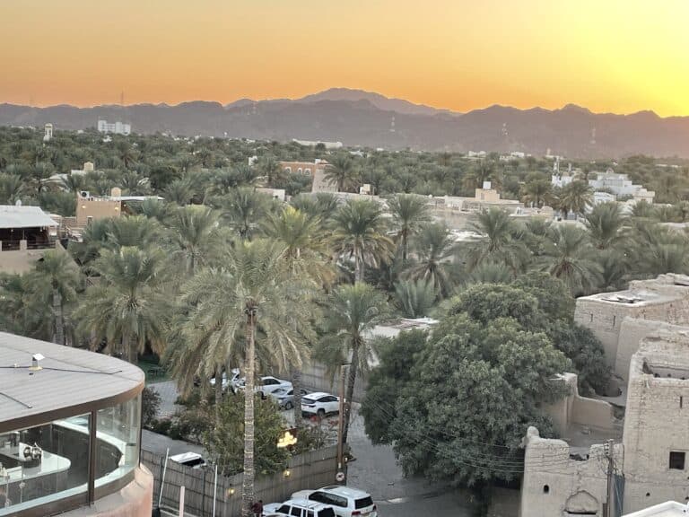 Aerial view of a lush, green oasis town (Nizwa) with numerous palm trees and tan buildings. Mountains are visible in the background under a golden sunset sky. Modern and traditional structures coexist in the landscape.