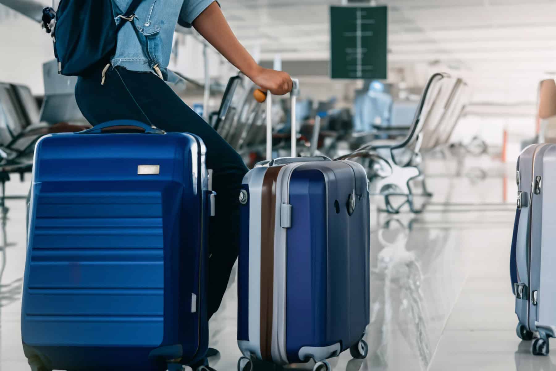 Person sitting on a large blue suitcase, holding a handle of another blue suitcase, in an airport terminal with chairs and a directional sign in the background.