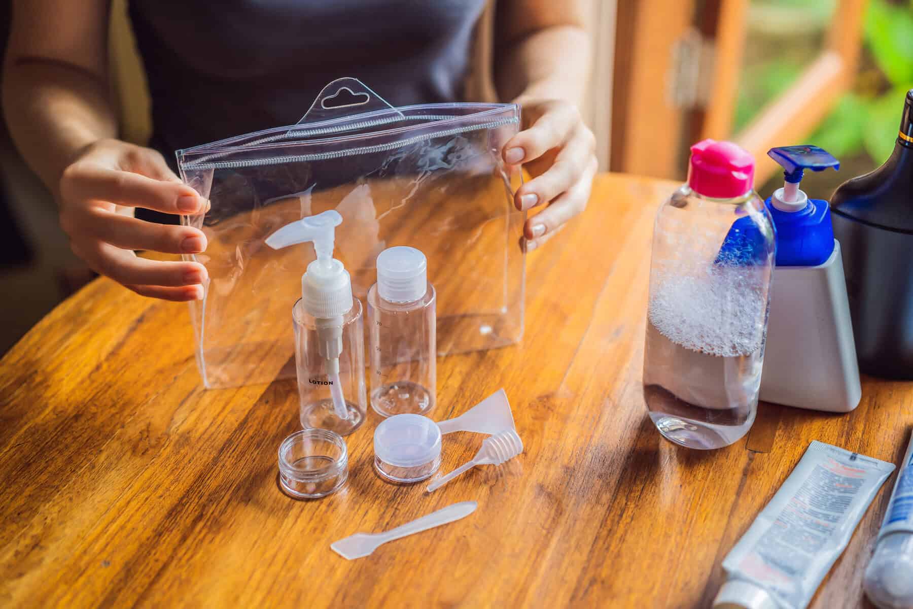 Person organizing travel-sized toiletry bottles and containers on a wooden table. A clear plastic bag holds several empty bottles with pumps and caps. Nearby are larger bottles with coloured caps, a small spatula, funnels, and a tube of cream.