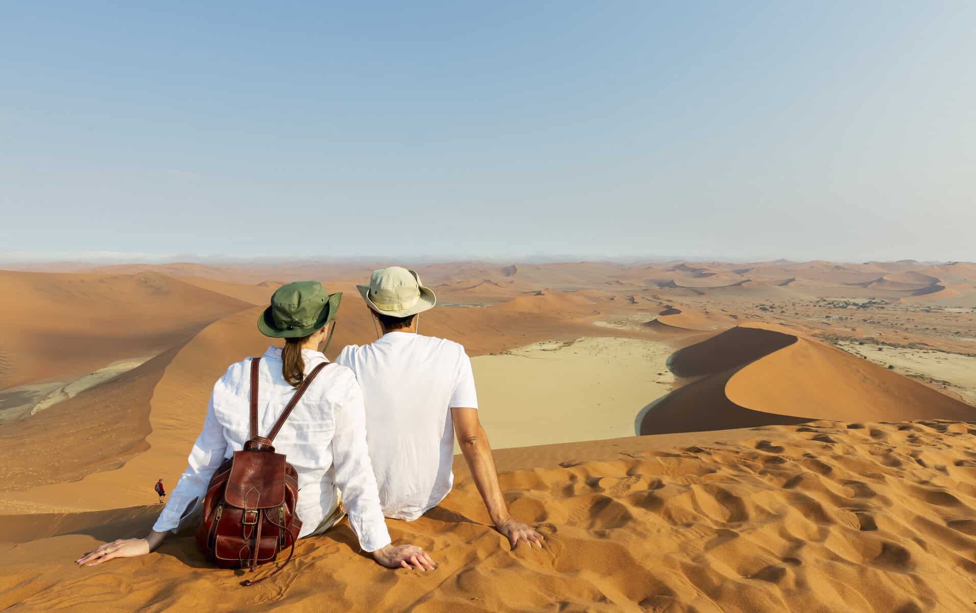 A couple with backpacks and sun hats sits on a sand dune, gazing at a vast desert landscape with rolling red dunes under a clear blue sky.