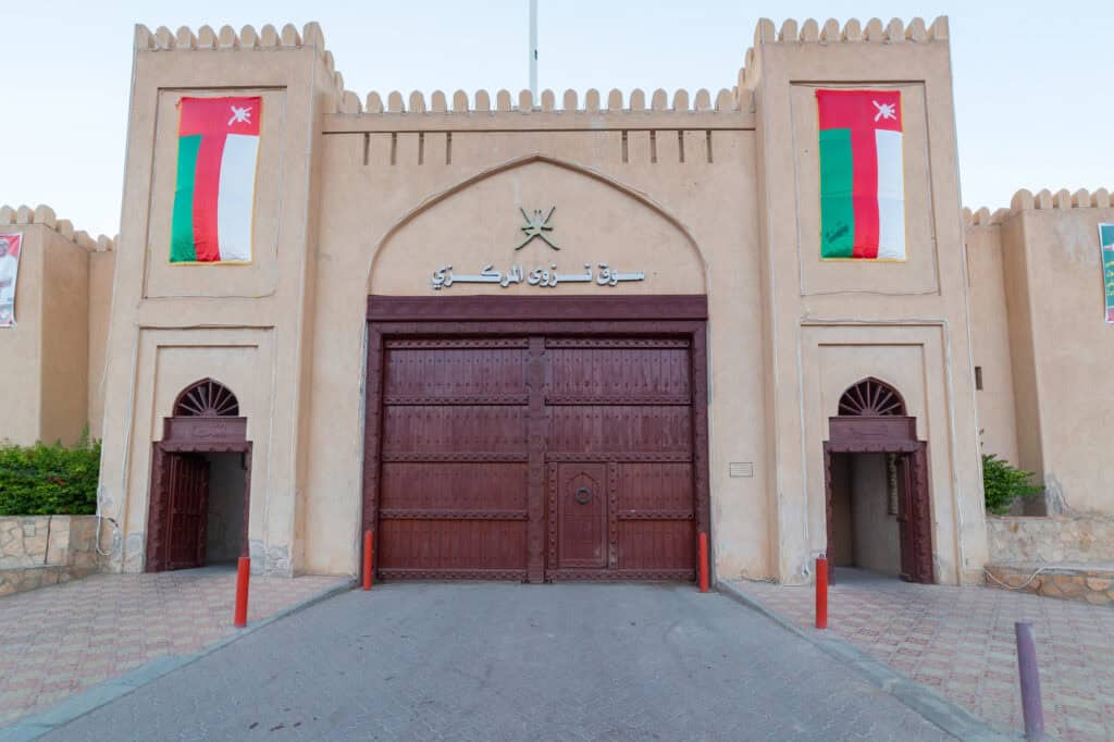 Main entrance of Nizwa Souq in Oman, showing the large wooden gate, Omani flags, and traditional fort-style architecture