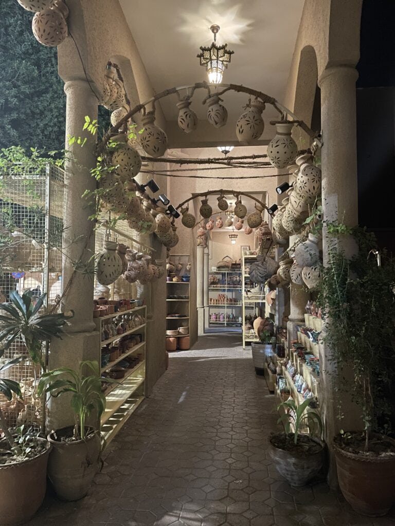 Early morning at Nizwa Souq pottery section, showing hanging clay lanterns, shelves of pottery, and soft shadows across the tiled walkway.