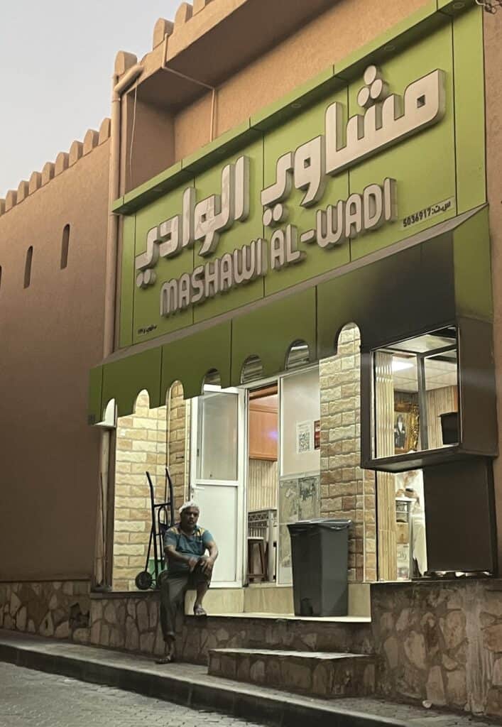 Exterior of Mashawi Al Wadi, a small mishkak cafe in Nizwa located opposite the post office, with a man sitting on the steps outside