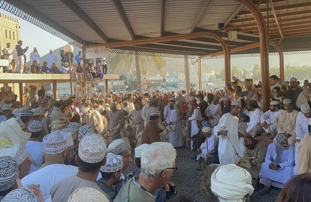 Crowds gather under a covered pavilion at the Nizwa Goat Market, with men leading goats through the ring