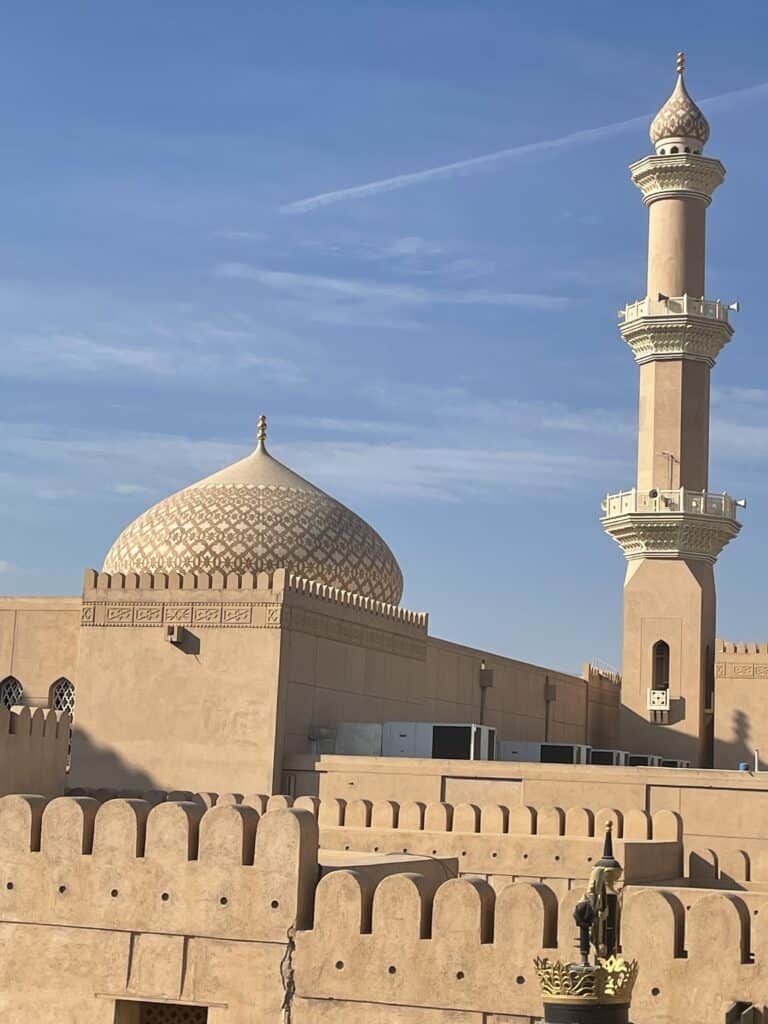 Al Qala’a Mosque near Nizwa Fort, showing its patterned dome and tall minaret rising above sand-coloured fort walls under a clear blue sky.