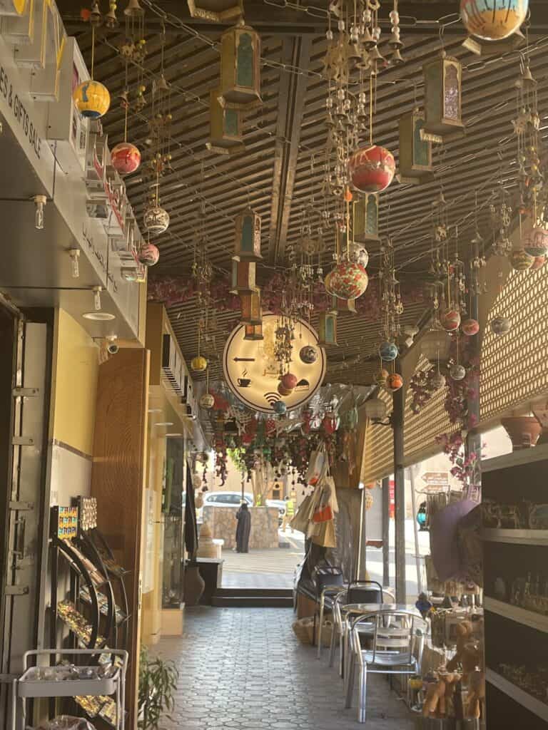 Covered walkway lined with small shops on the street leading to Nizwa Fort, decorated with hanging lanterns, bells, and colourful ornaments.